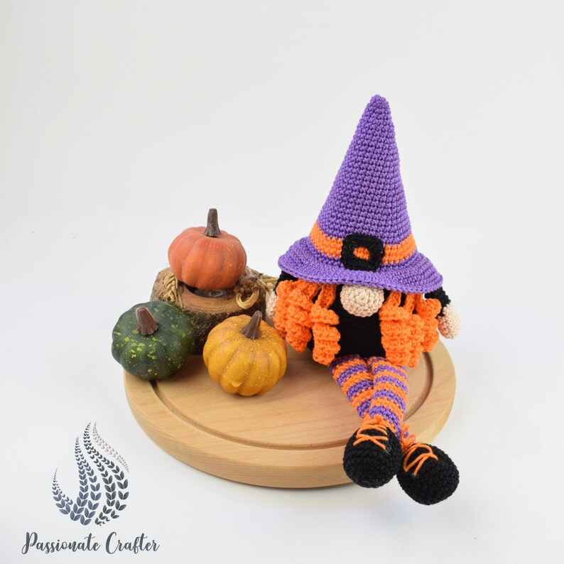 May include: A crocheted witch gnome wearing a purple hat with an orange and black striped outfit, sitting on a wooden surface with three small pumpkins.