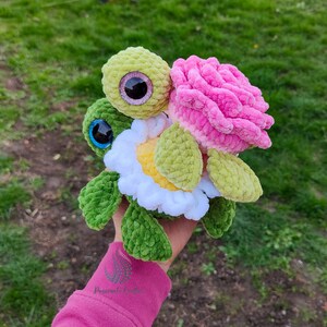 May include: A crocheted sea turtle toy with a pink flower on its back. The turtle is green and yellow and has large blue eyes. The flower is pink and white.