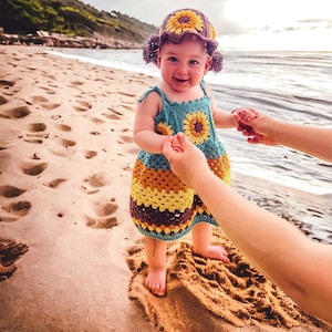 K&ouml;nnte beinhalten: Ein kleines M&auml;dchen tr&auml;gt einen geh&auml;kelten Sonnenblumenhut und ein Kleid am Sandstrand. Das Kleid hat eine blaue, gelbe und braune Farbgebung.