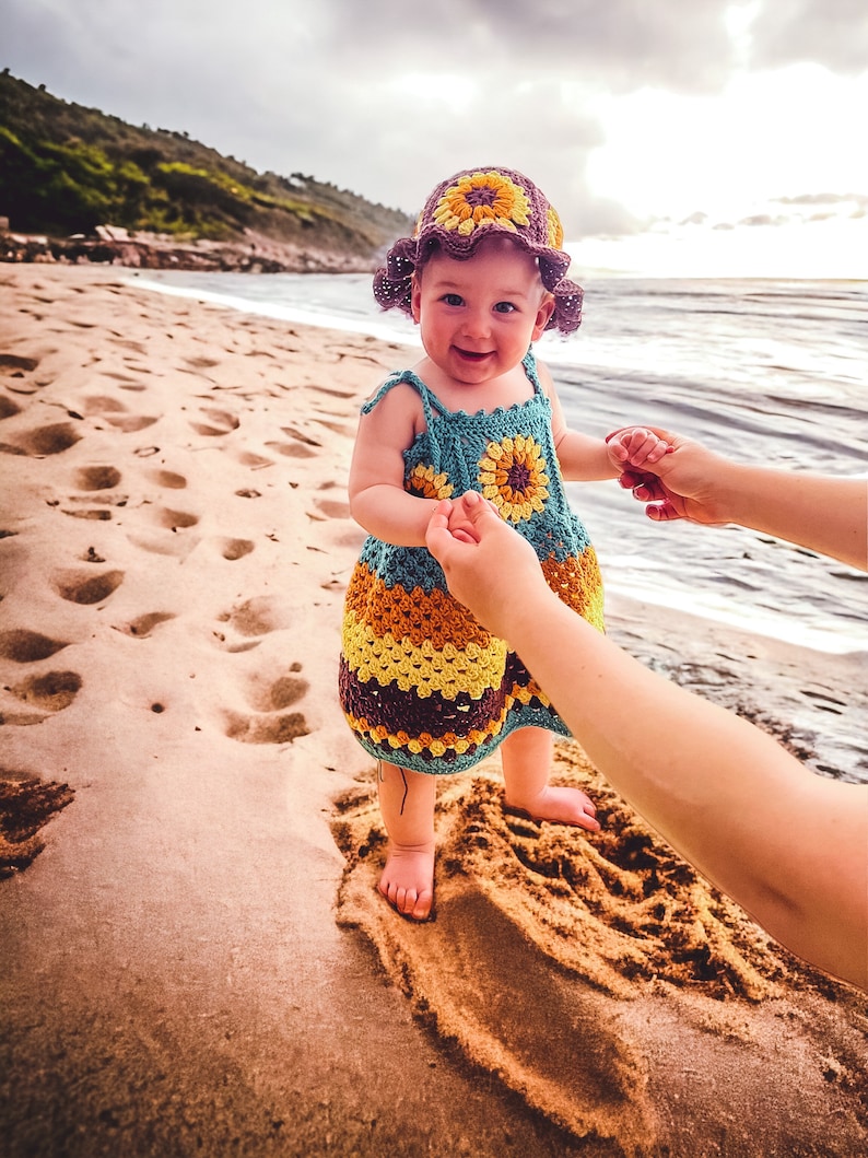 K&ouml;nnte beinhalten: Ein kleines M&auml;dchen tr&auml;gt einen geh&auml;kelten Sonnenblumenhut und ein Kleid am Sandstrand. Das Kleid hat eine blaue, gelbe und braune Farbgebung.
