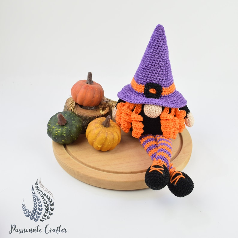 May include: A purple and orange crocheted witch gnome with a black hat and a black and orange striped outfit sitting on a wooden surface with three small pumpkins.