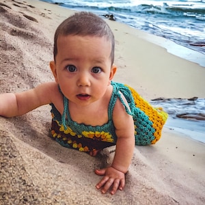 K&ouml;nnte beinhalten: Ein Baby in einem geh&auml;kelten Kleid mit Sonnenblumenmuster krabbelt an einem Sandstrand. Das Kleid ist t&uuml;rkis, gelb und braun.