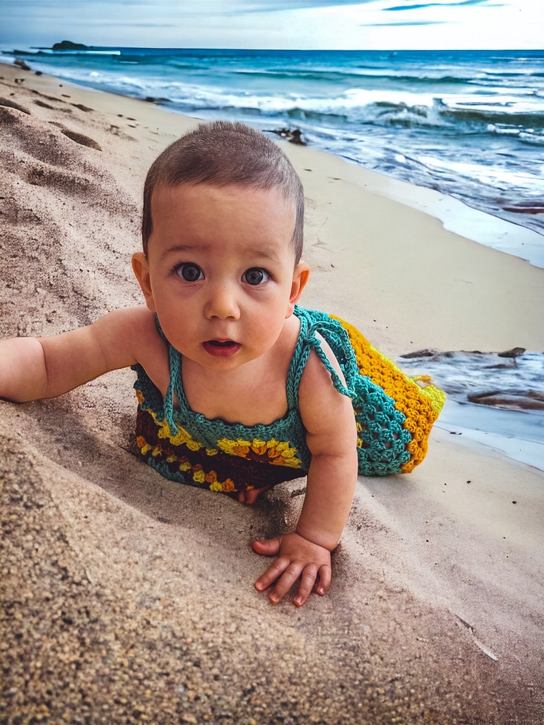 K&ouml;nnte beinhalten: Ein Baby in einem geh&auml;kelten Kleid mit Sonnenblumenmuster krabbelt an einem Sandstrand. Das Kleid ist t&uuml;rkis, gelb und braun.