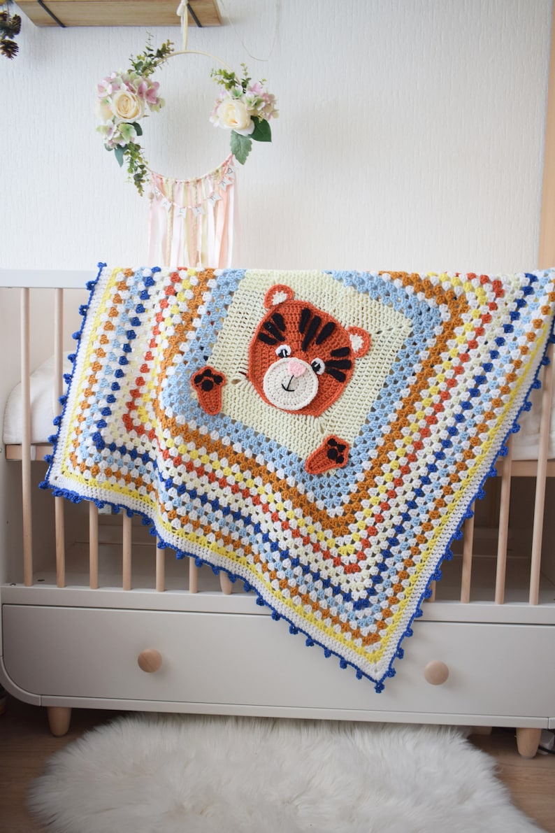 Peut inclure: Une couverture de b&eacute;b&eacute; en crochet color&eacute;e avec un visage de tigre au centre. La couverture est faite de fil bleu, jaune, orange et blanc. La couverture est drap&eacute; sur un lit b&eacute;b&eacute; blanc avec un cadre en bois.