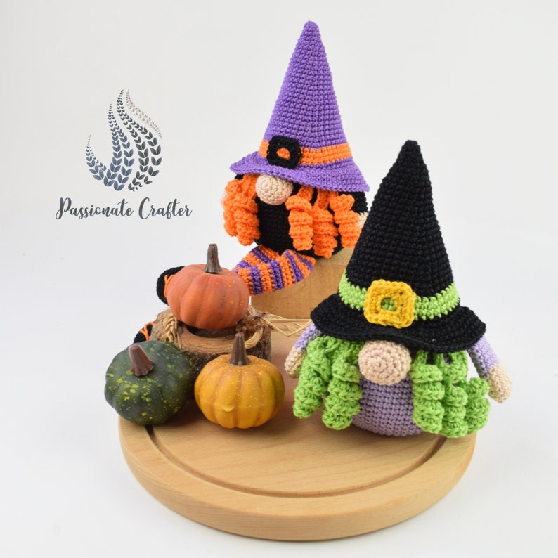 May include: Two crocheted Halloween gnomes with witch hats, one purple and one black, sitting on a wooden surface with small pumpkins.
