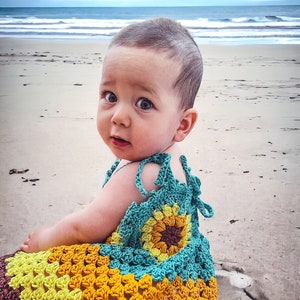 K&ouml;nnte beinhalten: Ein kleines M&auml;dchen tr&auml;gt ein geh&auml;keltes Kleid mit einem Sonnenblumenmuster in Braun-, Gelb- und T&uuml;rkist&ouml;nen. Das Kleid hat einen geh&auml;kelten Riemen, der sich im R&uuml;cken binden l&auml;sst. Das Baby sitzt auf einem Sandstrand mit dem Meer im Hintergrund.