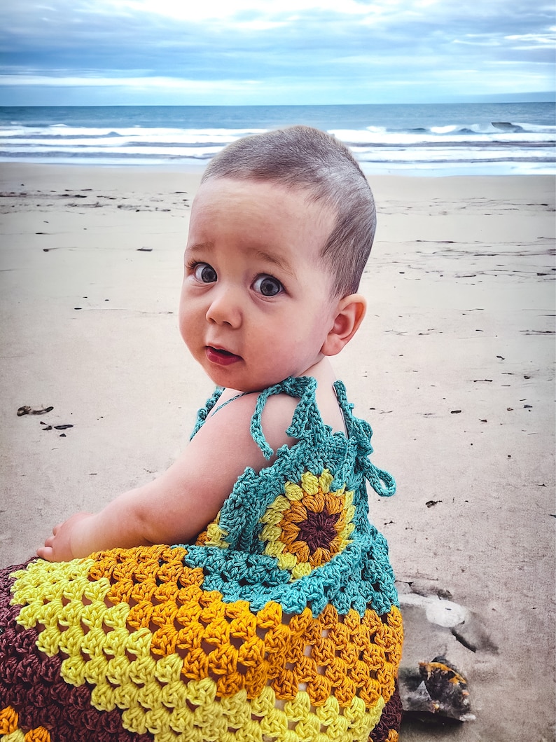 K&ouml;nnte beinhalten: Ein kleines M&auml;dchen tr&auml;gt ein geh&auml;keltes Kleid mit einem Sonnenblumenmuster in Braun-, Gelb- und T&uuml;rkist&ouml;nen. Das Kleid hat einen geh&auml;kelten Riemen, der sich im R&uuml;cken binden l&auml;sst. Das Baby sitzt auf einem Sandstrand mit dem Meer im Hintergrund.