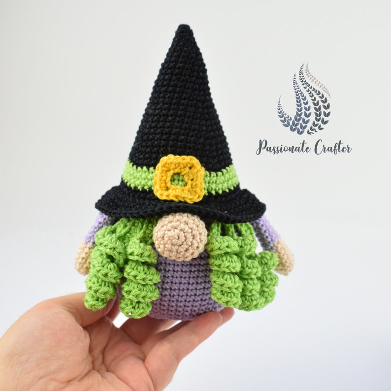May include: A crocheted witch gnome with a black pointed hat, green and purple hair, and a yellow buckle.
