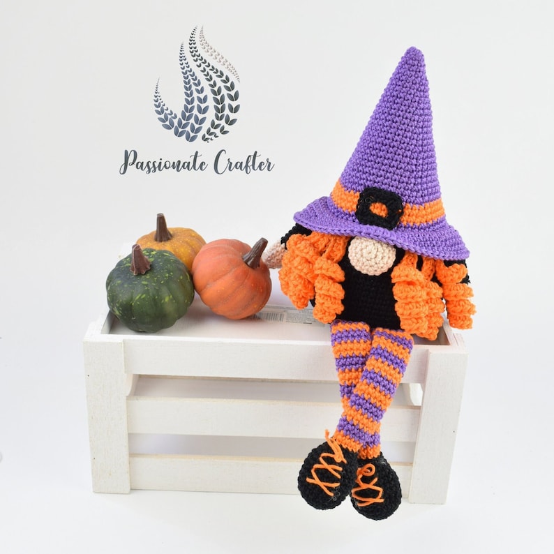 May include: A purple and orange crocheted witch gnome with a black hat and long legs. The gnome is sitting on a white crate with three small pumpkins in front of it.