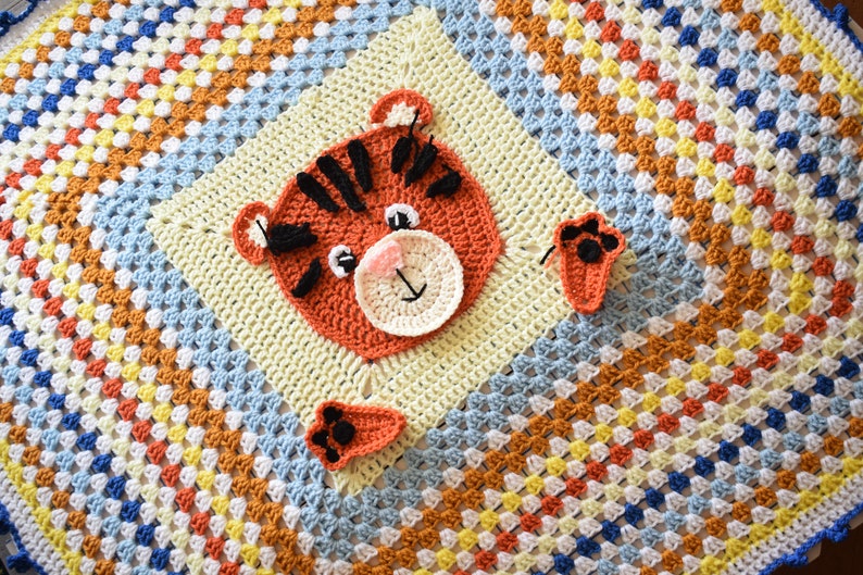 Peut inclure: Une couverture pour b&eacute;b&eacute; en crochet avec un motif de carr&eacute;s de grand-m&egrave;re color&eacute;s. La couverture pr&eacute;sente un visage de tigre en crochet et deux empreintes de pattes.