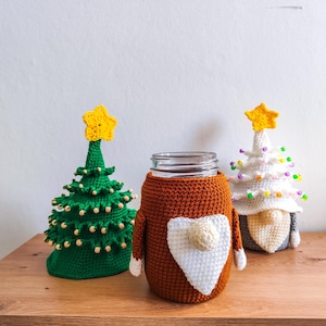 May include: Three crocheted Christmas tree ornaments. One is a green tree with a yellow star on top, another is a white tree with a yellow star on top, and the third is a brown gnome-shaped jar cover with a white beard and a white star on top.