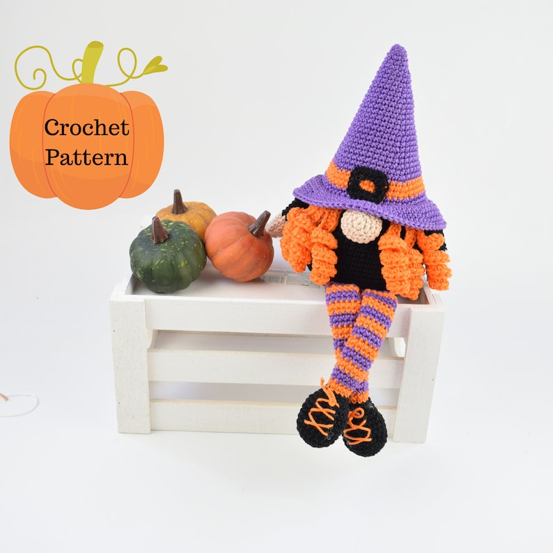 May include: A crocheted witch gnome with a purple hat, orange hair, and black and orange striped legs. The gnome is sitting on a white crate with three small pumpkins in front of it.