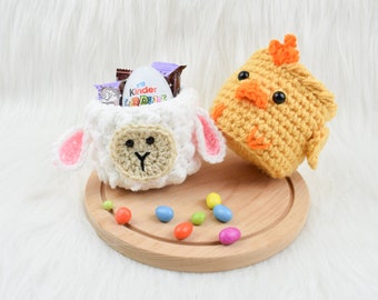 May include: Crocheted Easter-themed containers shaped like a lamb and a chick. The white lamb has pink ears and a tan face, while the yellow chick has an orange comb. The lamb holds a Kinder Joy egg and candies. The items are on a wooden tray.