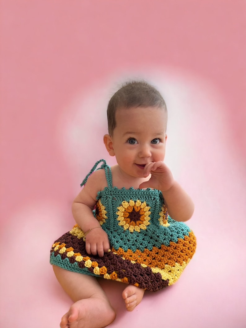 K&ouml;nnte beinhalten: Ein kleines M&auml;dchen tr&auml;gt ein geh&auml;keltes Kleid mit einem mehrfarbigen Muster aus Braun, Gelb und Gr&uuml;n. Das Kleid hat ein quadratisches Design mit einer Blume in der Mitte.