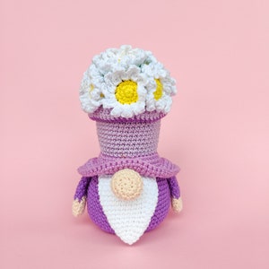 May include: A crocheted gnome wearing a purple hat with a white flower crown. The gnome has a white beard and a purple body.