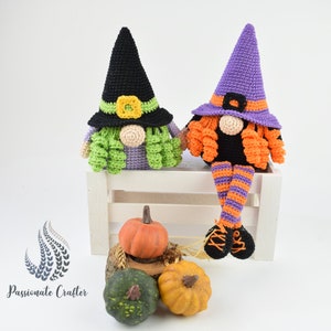 May include: Two crocheted witch gnomes sitting on a white wooden bench. The gnome on the left is wearing a black witch hat with a green and yellow flower accent. The gnome on the right is wearing a purple witch hat with orange and purple striped legs. Both gnomes have orange hair and are sitting on a white wooden bench with pumpkins in front of them.