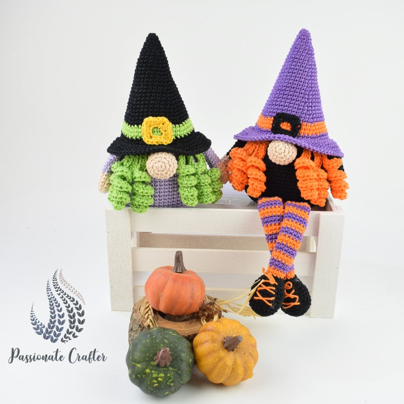 May include: Two crocheted witch gnomes sitting on a white wooden bench. The gnome on the left is wearing a black witch hat with a green and yellow flower accent. The gnome on the right is wearing a purple witch hat with orange and purple striped legs. Both gnomes have orange hair and are sitting on a white wooden bench with pumpkins in front of them.