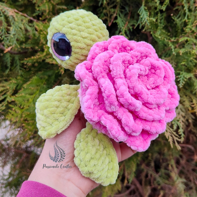 May include: A crocheted green turtle with a pink rose on its back. The turtle has a large, black eye and a small, white mouth. The rose is made of soft, fluffy yarn.