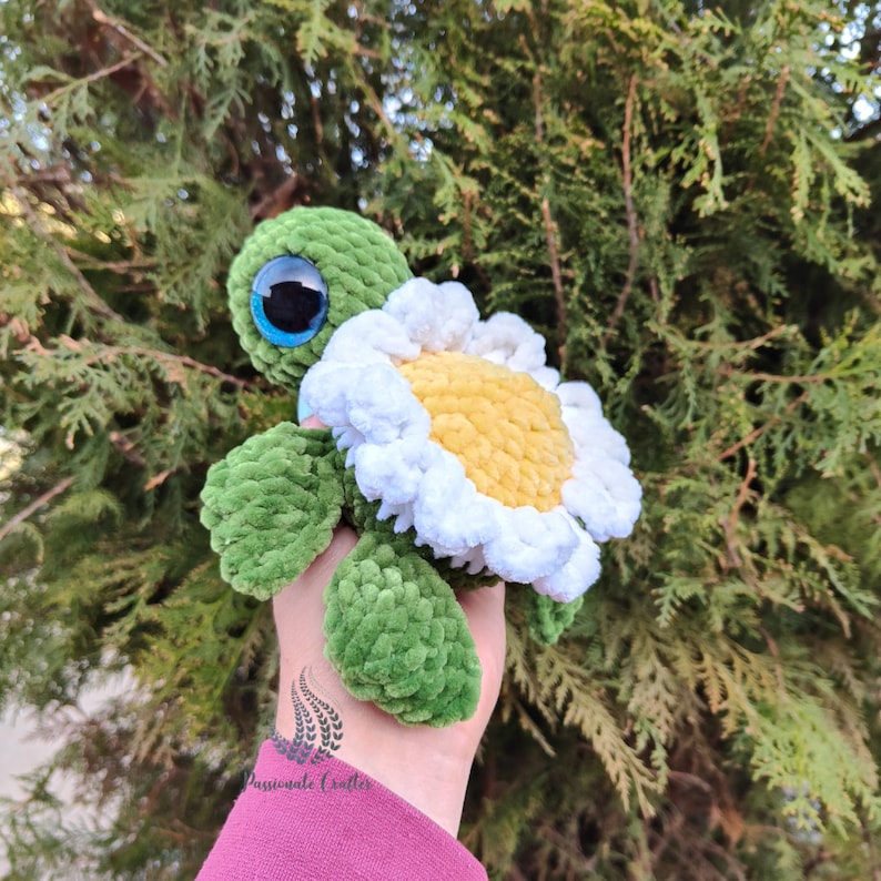 May include: Green crocheted turtle with a white and yellow crocheted flower on its back. The turtle has large blue eyes.