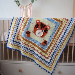 Peut inclure: Une couverture de b&eacute;b&eacute; en crochet color&eacute;e avec un visage de tigre au centre. La couverture est faite de plusieurs couleurs, dont le bleu, le jaune, l'orange et le blanc. La couverture est drap&eacute;e sur un lit b&eacute;b&eacute; blanc avec un cadre en bois.