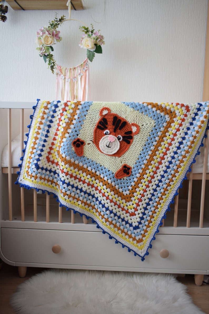 Peut inclure: Une couverture de b&eacute;b&eacute; en crochet color&eacute;e avec un visage de tigre au centre. La couverture est faite de plusieurs couleurs, dont le bleu, le jaune, l'orange et le blanc. La couverture est drap&eacute;e sur un lit b&eacute;b&eacute; blanc avec un cadre en bois.