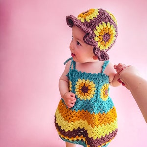 K&ouml;nnte beinhalten: Ein kleines M&auml;dchen tr&auml;gt ein geh&auml;keltes Sonnenblumenkleid und einen Hut in Braun-, Gelb- und T&uuml;rkist&ouml;nen. Das Kleid hat ein mehrschichtiges Design mit einem gewellten Saum. Das Baby steht auf einem rosa Hintergrund.