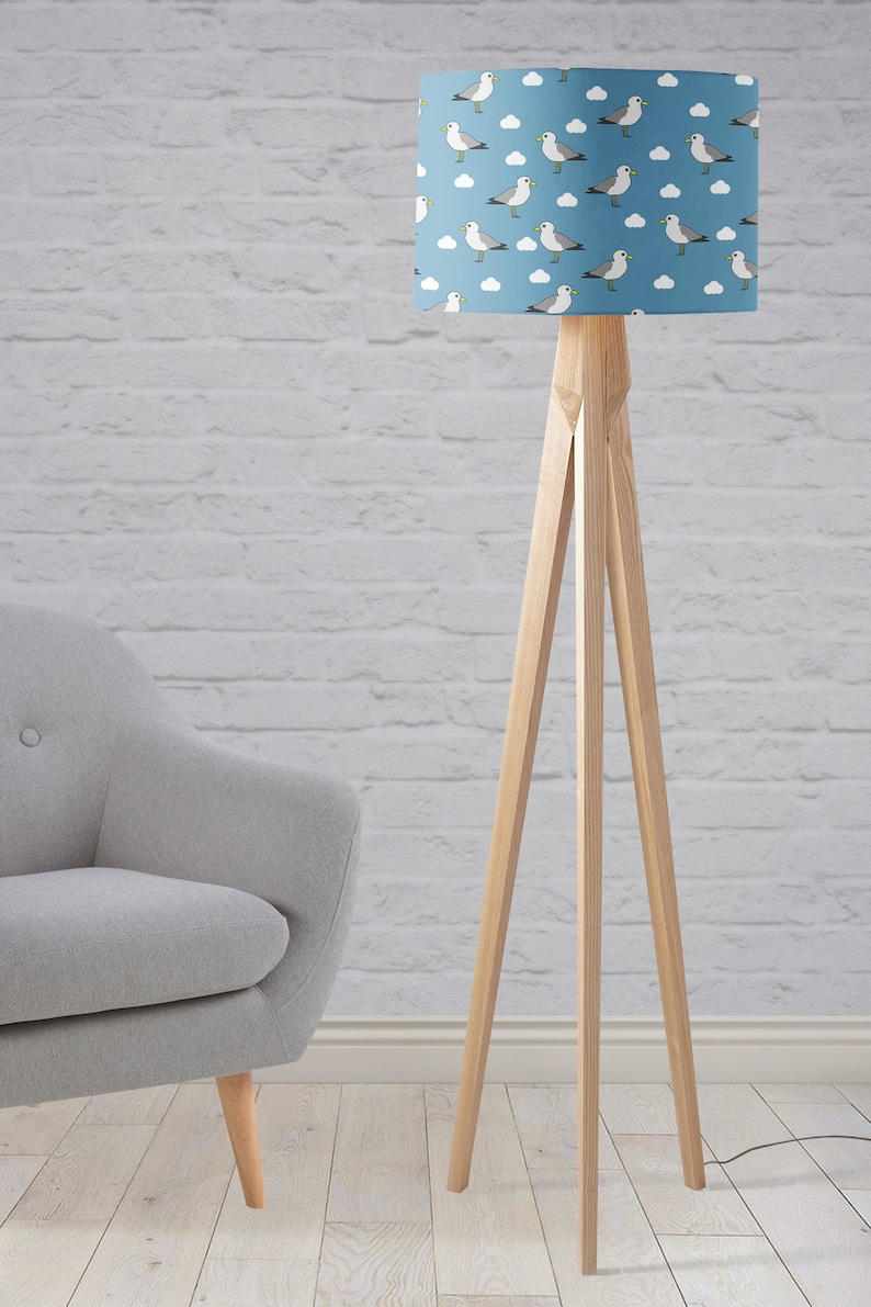 May include: A wooden floor lamp with a blue lampshade featuring a white seagull and cloud pattern. The lamp has a tripod base.