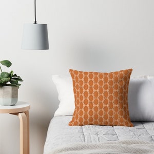 Op de afbeelding: Een kussen met een geometrisch patroon in wit op een oranje-bruine achtergrond. Het kussen ligt op een bed met een wit dekbed en een wit kussen.
