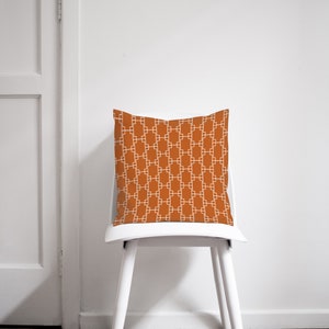 Op de afbeelding: Een witte stoel met een oranjegeel kussen met een wit geometrisch patroon. Het kussen is vierkant en heeft een eenvoudig ontwerp.