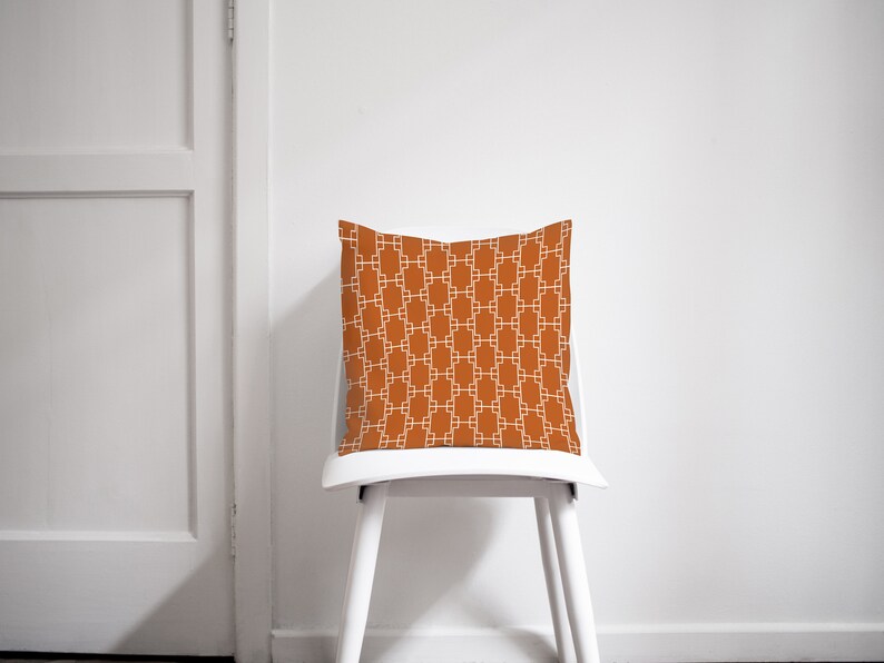 Op de afbeelding: Een witte stoel met een oranjegeel kussen met een wit geometrisch patroon. Het kussen is vierkant en heeft een eenvoudig ontwerp.
