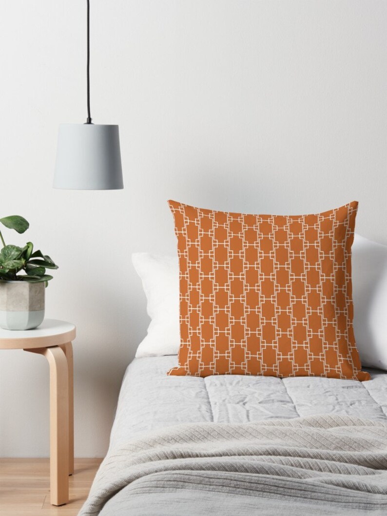 Op de afbeelding: Een kussen met een wit geometrisch patroon op een oranje-bruine achtergrond. Het kussen ligt op een bed met een wit dekbedovertrek en een witte deken.