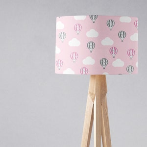 May include: A pink lampshade with a pattern of gray and pink hot air balloons and white clouds. The lampshade is on a wooden tripod stand.