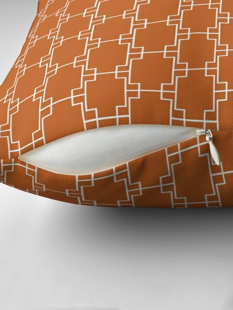 Op de afbeelding: Een kussen met een geometrisch patroon in wit op een oranje-bruine achtergrond. Het kussen heeft een ritssluiting.
