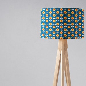 May include: A floor lamp with a blue lampshade featuring a repeating pattern of yellow flowers with red centers. The lampshade is cylindrical and the lamp has a wooden tripod base.