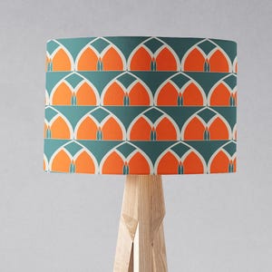Op de afbeelding: Een lampenkap met een geometrisch patroon in teal en oranje. De lampenkap staat op een houten lampenvoet met drie poten.
