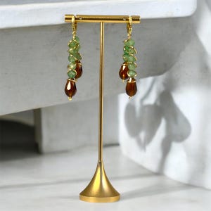 Pode incluir: Um par de brincos pendurados com uma armação de metal dourado. Os brincos apresentam uma combinação de pequenas contas verdes claras e contas maiores, em forma de gota, castanho escuro. Os brincos são exibidos em um suporte dourado.