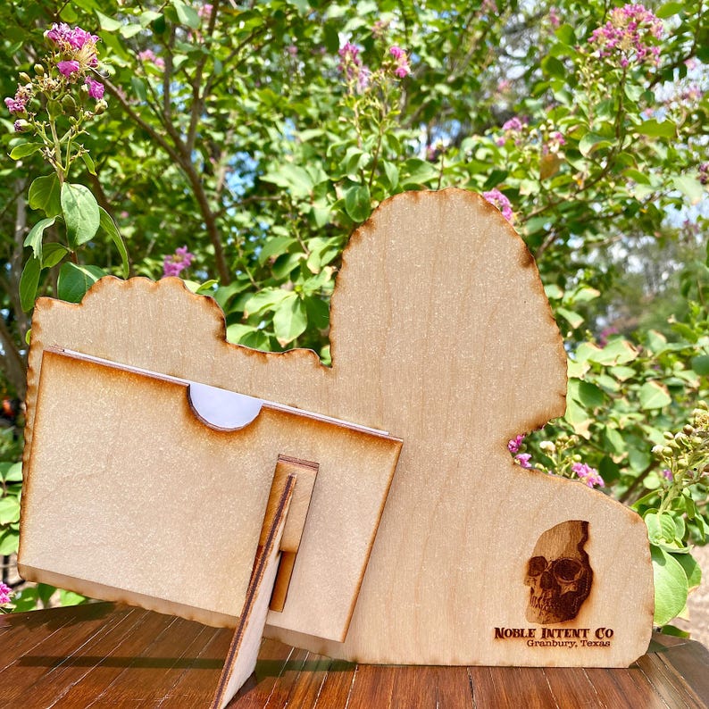 K&ouml;nnte beinhalten: Holzschnitt des Bundesstaates Texas mit einem kleinen St&auml;nder zum Ausstellen von Fotos oder Karten. Der Ausschnitt hat ein Totenkopfdesign und den Text "NOBLE INTENT CO. Granbury, Texas".
