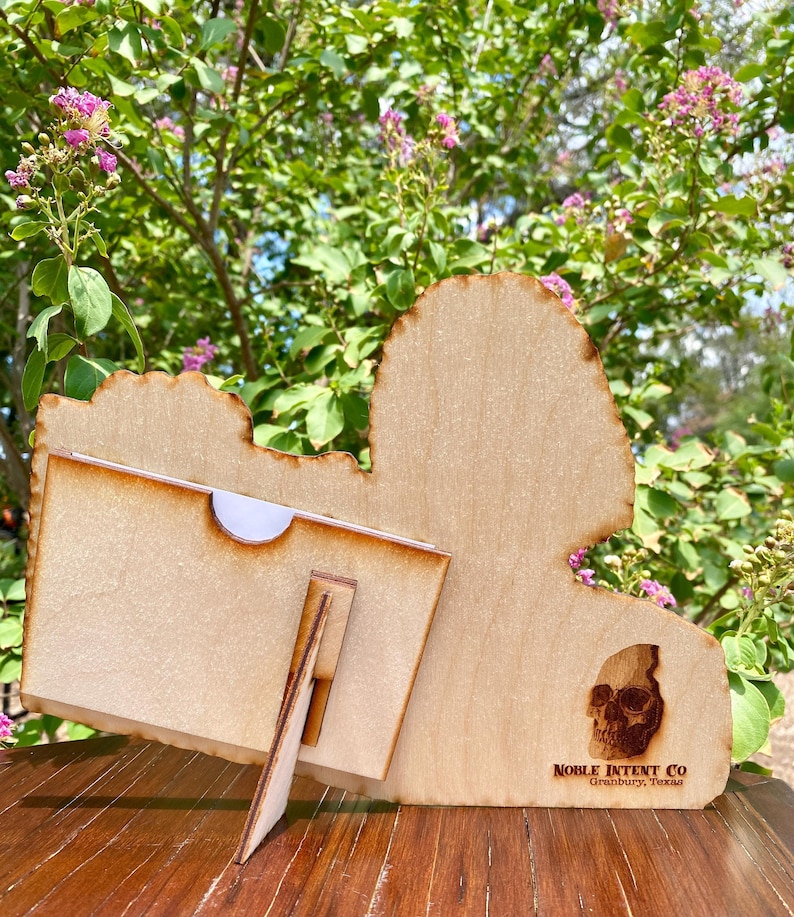 K&ouml;nnte beinhalten: Holzschnitt eines Sch&auml;dels mit einem St&auml;nder. Der Schnitt ist braun und hat einen verwitterten Look. Der St&auml;nder ist aus Holz und braun lackiert. Der Schnitt hat den Text "NOBLE INTENT CO. Granbury, Texas" unten.