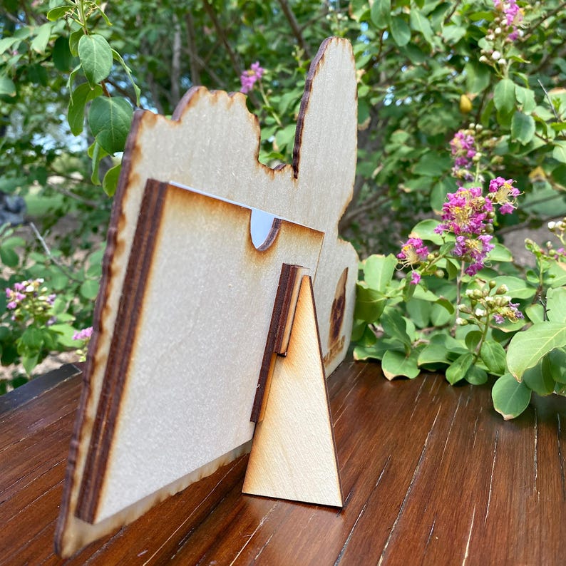K&ouml;nnte beinhalten: Ein Bilderrahmen aus Holz mit einem ausgeschnittenen Design. Der Rahmen hat einen St&auml;nder und ist aus hellem braunem Holz.