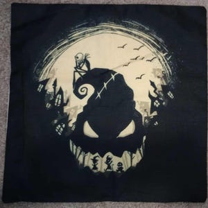 May include: Black decorative pillow featuring a spooky design. The design includes a large, grinning face with a hat, a full moon, and silhouettes of buildings and characters. A skeleton figure sits on a hill. The pillow is square.