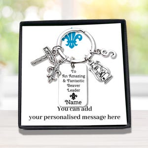 May include: A silver-toned metal keyring featuring a circular ring with four charms: a signpost labelled CAMPING, a fleur-de-lis, a lantern, and the letter S. A rectangular tag is engraved with To An Amazing & Fantastic Beaver Leader and Name.