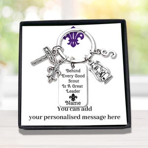 May include: A silver-coloured metal keyring featuring a circular ring with four charms: a signpost labelled CAMPING, a fleur-de-lis, a lantern, and the letter S. A rectangular tag hangs from the ring with the text Behind Every Good Scout Is A Great Leader.