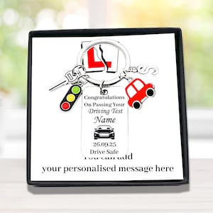 May include: A silver metal keyring featuring a rectangular tag with the text Congratulations On Passing Your Driving Test Name, a car icon, the date 26.09.25, and Drive Safe. Charms include a traffic light, a red car, and a letter S.