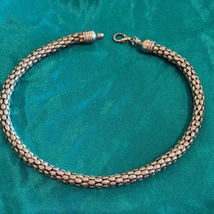 Può includere: Una collana girocollo in metallo color argento caratterizzata da un design a catena a maglia intrecciata flessibile. La collana presenta un terminale cilindrico su un lato e una chiusura a moschettone sull'altro per fissare la forma circolare.