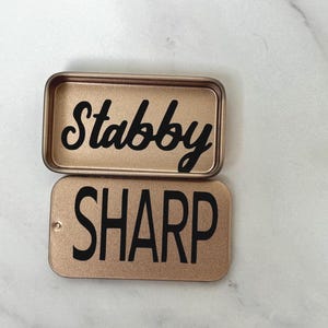 May include: Two rectangular metallic gold tins with rounded corners. The top tin is open and features the word Stabby in black cursive script. The bottom tin is closed and features the word SHARP in black sans-serif capital letters.