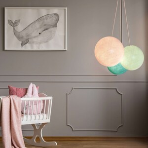 May include: A white crib with a pink blanket and pillows, a whale print on the wall, and a hanging light fixture with three round, pastel-colored shades.