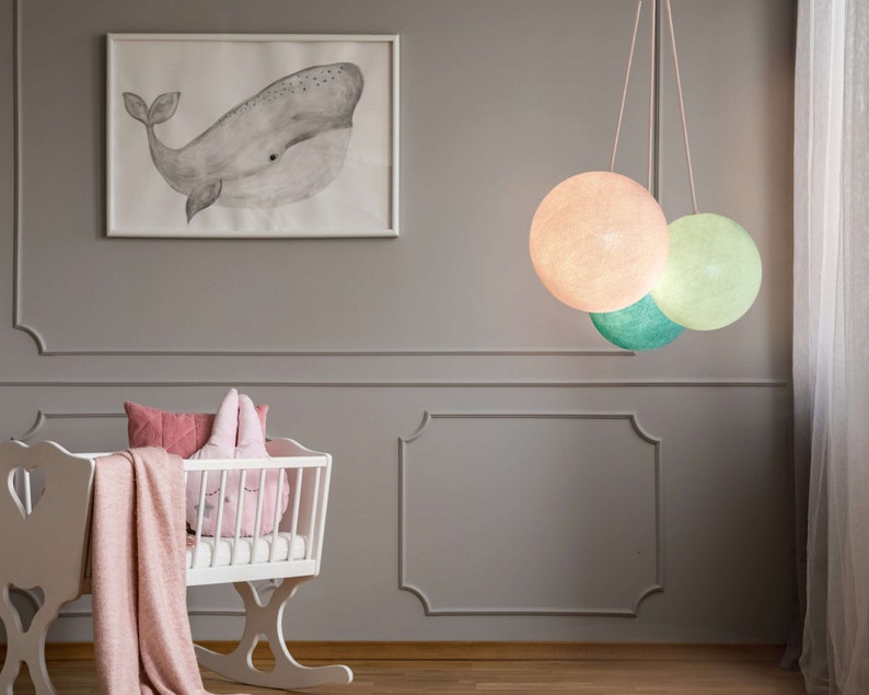 May include: A white crib with a pink blanket and pillows, a whale print on the wall, and a hanging light fixture with three round, pastel-colored shades.