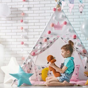 May include: A young girl sits inside a white and pink teepee tent with a stuffed animal. The teepee is decorated with stars and pom-poms. There are balloons and a white brick wall in the background.