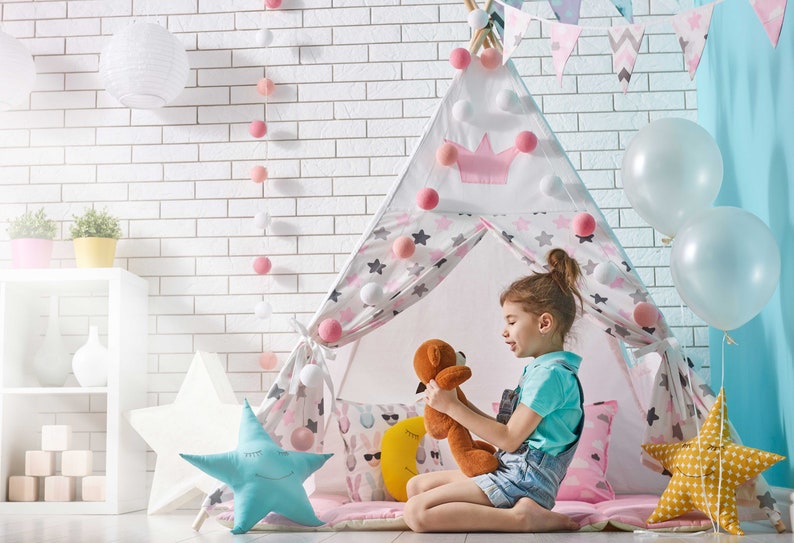 May include: A young girl sits inside a white and pink teepee tent with a stuffed animal. The teepee is decorated with stars and pom-poms. There are balloons and a white brick wall in the background.