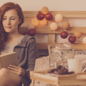 May include: A woman sits in bed reading a book. The bed is decorated with string lights and pillows. There is a tray with a mug and a glass jar on a small table next to the bed.